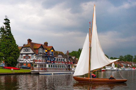 The Swan Inn at Horning on the River Bure is part of the Norfolk Broads. Going past is a small sailing boat.のeditorial素材