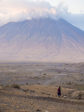 Maasai warrior walks in front of the Ol Doinyo Lengai Mountain of God in the Maasai language, an active volcano in Arusha Region i the Northern Tanzania, Africa, at sunrise.のeditorial素材