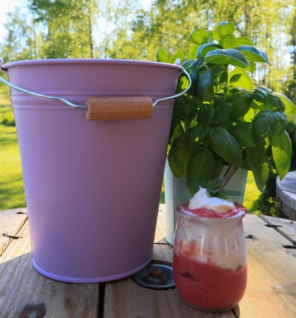 appetizing homemade strawberry dessert with whipped cream in a small jar with a spoon standing on wooden brown boards against a green background and a purple pastel bucketの写真素材