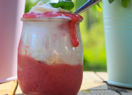 appetizing homemade strawberry dessert with whipped cream in a small jar with a spoon standing on wooden brown boards against a green background with colouret pots aroundの写真素材
