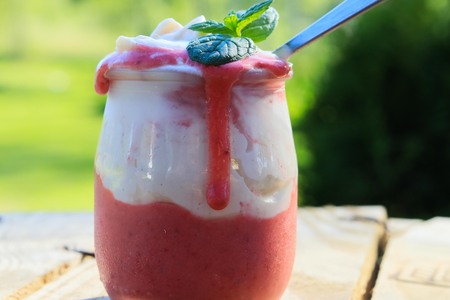 appetizing homemade strawberry dessert with whipped cream in a small jar with a spoon standing on wooden brown boards against a green backgroundの写真素材