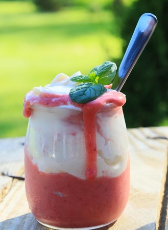 appetizing homemade strawberry dessert with whipped cream in a small jar with a spoon standing on wooden brown boards against a green backgroundの写真素材