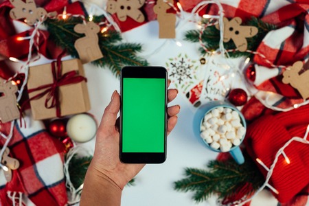Flat lay of Woman hands holding black smartphone with green screen on white table with beautiful Christmas decorations. Mockup for app presentation. Chroma keyの写真素材