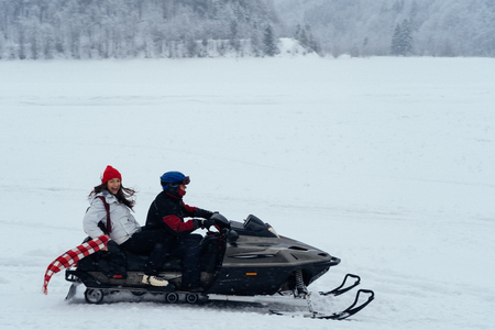 Side view of man and woman riding fast on a snowmobile on the frozen lake in the mountains with the scenic view. Pine trees covered with snowの写真素材