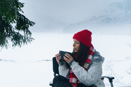 Beautiful woman sitting near fire with a cup of tea in the winter forest with scenic mountain view. Tourist camp with campfire. Camping and hiking, winter adventure, nature landscape. Warming upの写真素材