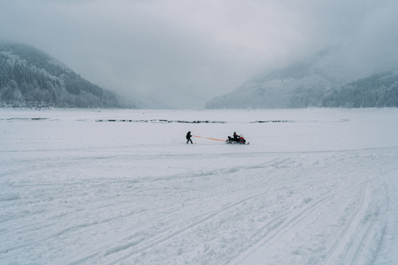 Man snowboarding holding a rope towed behind a snowmobile on the frozen lake between beautiful snowy...