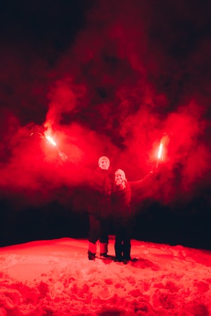Couple of man and woman in winter clothes standing on the top of the hill with the red fire torches at night. Red smoke. Fire show. Valentines dayの写真素材