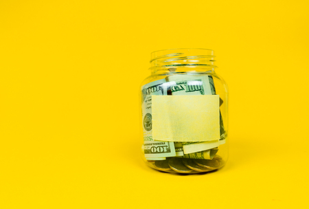 Glass money jar with coins and paper dollars with sticker for your text, copy space. Isolated on yellow backgroundの写真素材