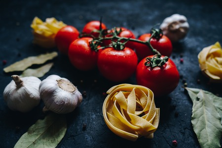 Fresh organic tomatoes and tagliatelle with garlic and herbs on a black background. Ingredients for pastaの写真素材