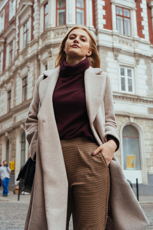 Attractive young blonde woman in casual coat smiling at the camera on the city street. Beautiful architecture building on backgroundの写真素材
