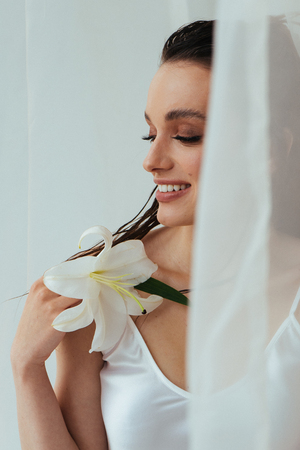 Beauty portrait of young brunette woman with beautiful make up, tan sking holding lily flower while posing on white background. Skincare cosmetics, Wellnessの写真素材