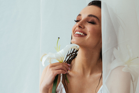 Portrait of beautiful sensual brunette woman with tan skin, make up holding lily flower and looking through the curtain. Skincare cosmetics. Fresh, natural skin treatment.の写真素材