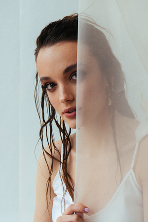 Beautiful young woman with nude make up and long wet brunette hair smiling while looking through the curtain. White background. Beauty portrait. Skincare treatmentの写真素材