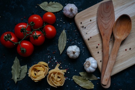 Fresh raw tomatoes, garlic, pasta and wooden spatula and a spoon on a wooden cutting board isolated on black table background. Top view. Pasta ingredientsの写真素材
