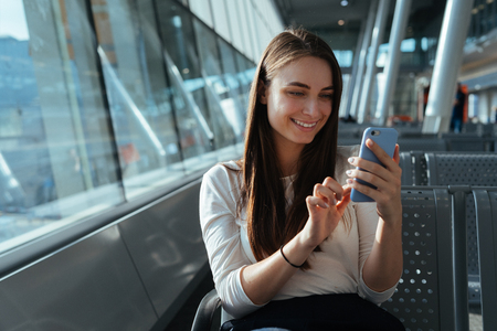 Young beautiful woman sitting at the gate and using smartphone while waiting for a flight at the airport. Travel concept. Passenger girl chatting and smiling in terminal departure lounge.の写真素材