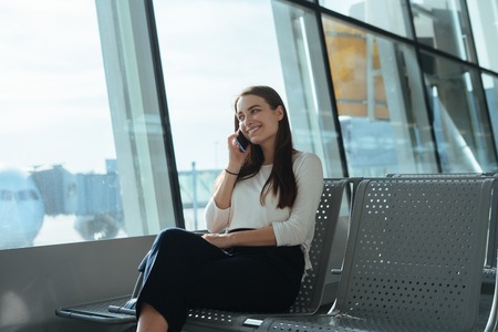 Young smiling woman sitting at the airport terminal departure lounge and talking on the cell phone while waiting for a flight. Travel concept. Passenger girl having a conversation on mobile phone.の写真素材