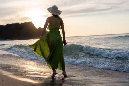 Beautiful woman in green dress and hat walking on the beach at sunsetの写真素材