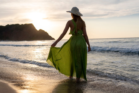 Beautiful young woman in green dress and straw hat walking on the beach at sunsetの写真素材