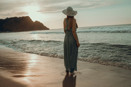 Young woman in straw hat and dress walking on beach at sunset.の写真素材