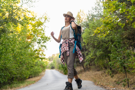 Young woman with a backpack and a hat is walking along a country road.の写真素材