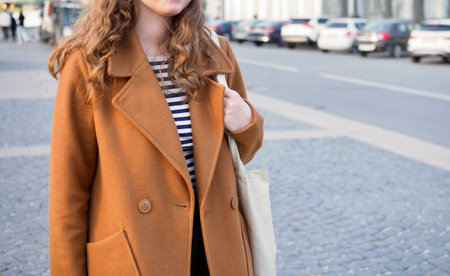stylish caucasian beautiful young girl with long curly hair walks around a European city in a brown coat and a white shopping bagの写真素材