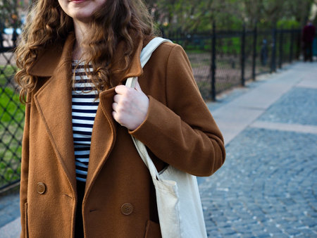 stylish caucasian beautiful young girl with long curly hair walks around a European city in a brown coat and a white shopping bagの写真素材