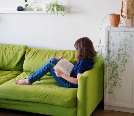 a little girl child is lying on the green couch and reading a book at homeの写真素材