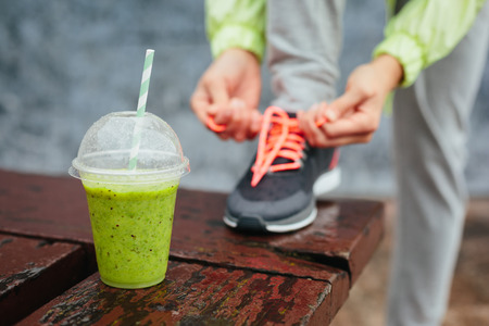Green detox smoothie cup and woman lacing running shoes before workout on rainy day Fitness and healthy lifestyle concept Green detox smoothie cup and woman lacing running shoes before workout on rainy day Fitness and healthy lifestyle concept