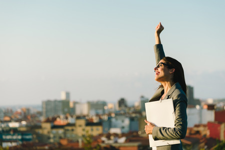 Successful young businesswoman raising arm celebrating business or job achievement towards city background. Professional happy woman walking outside. Successful young businesswoman raising arm celebrating business or job achievement towards city background. Professional happy woman walking outside.