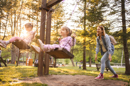 Mother is rocking her twin daughters on a swing in the parkの写真素材