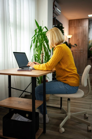 Young woman working at a cozy desk in a warm-toned room surrounded by greenery while typing on a laptop during the afternoonの写真素材