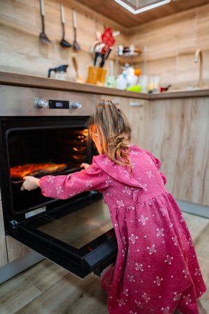 In a bright and warm kitchen, a young girl eagerly leans into the oven, her pink dress adorned with floral patternsの写真素材