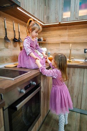 Sisters share laughter while playing dress-up in a cozy kitchen filled with warm wooden textures and soft lighting during a sunny afternoonの写真素材