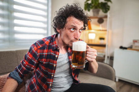 Young adult man sitting on sofa drinking beer at home