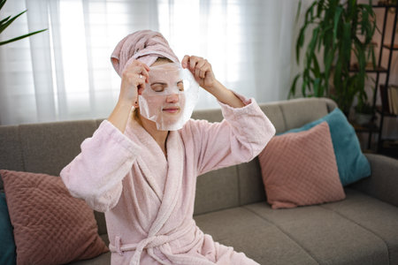 Woman applying a facial cosmetic mask at home sitting on the sofaの写真素材