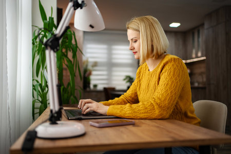 Focused woman in yellow sweater working on laptop at home desk with bright lamp and plants in a cozy atmosphereの写真素材