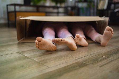 Two playful children hide inside a large cardboard box indoorsの写真素材