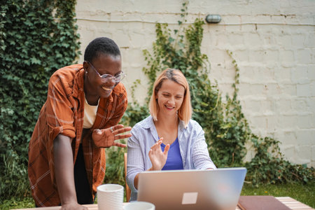 Two smiling female colleagues waving at laptop during video call in garden