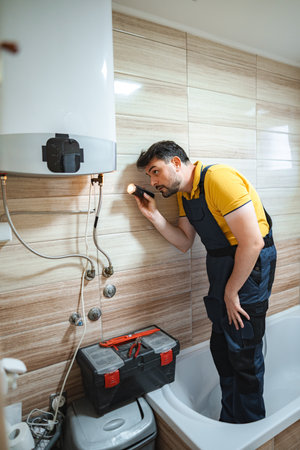 Plumber inspecting water heater in bathroom using flashlightの写真素材