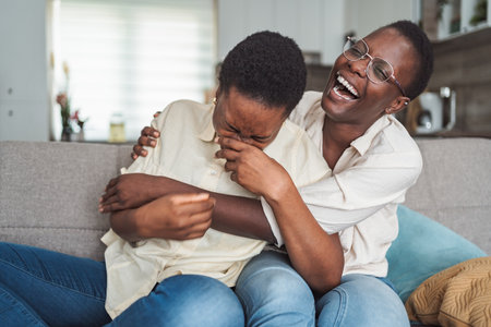 Two young black women laughing and hugging on the couch at homeの写真素材