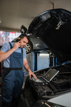 Car mechanic using laptop and talking on phone while inspecting vehicle in auto repair shopの写真素材