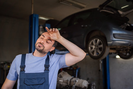 Exhausted mechanic wiping sweat from forehead in auto repair shopの写真素材