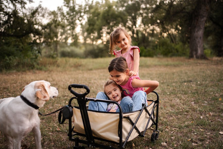Three little sisters having fun with their dog in a wagon in a parkの写真素材