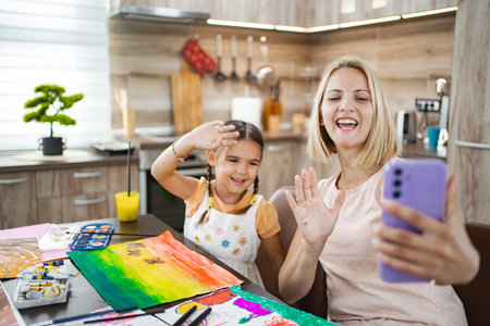 Mother and daughter enjoying art time while video chatting in a cozy kitchenの写真素材