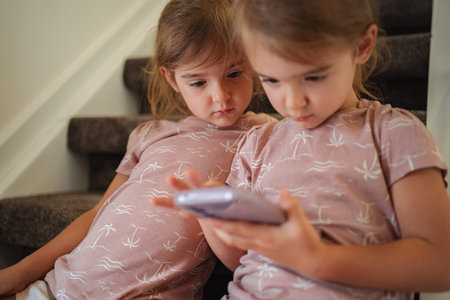 Two young sisters watching a smartphone sitting on stairs at home