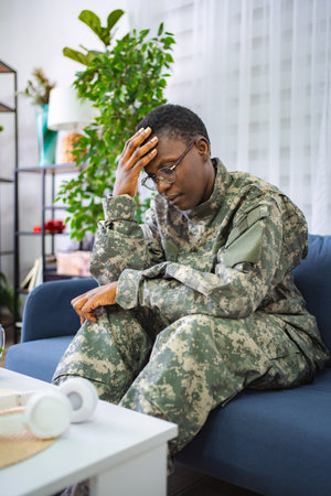 Depressed african american soldier sitting on sofa at home touching foreheadの写真素材