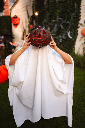 Child wearing ghost costume holding jack-o-lantern during Halloween party in the backyardの写真素材