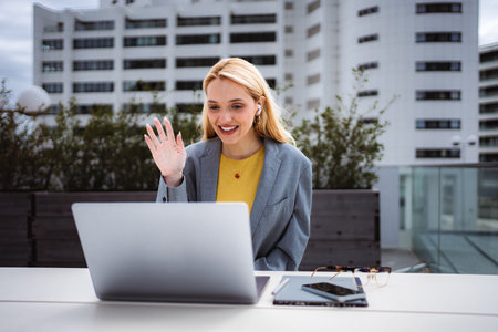 Young woman waving to computer during video call on urban rooftopの写真素材