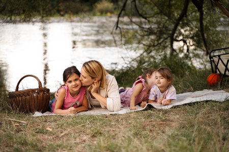 Mother kissing daughter during picnic by the river with twin sistersの写真素材