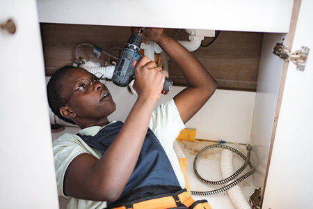 Young female plumber using electric screwdriver beneath kitchen sinkの写真素材